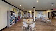 This single-floor activity and creative space is designed to foster engagement, learning, and artistic expression. The main open area is generously sized, featuring durable wood-look flooring, recessed lighting, and dark crown molding that frames the cream-colored walls. Multiple folding tables with chairs are arranged throughout the room, accommodating various group activities simultaneously.

A prominent feature wall showcases colorful open-shelf storage units filled with art supplies, books, and decorative items, alongside motivational signage including a bold "LOVE" display and a "CREATE" station equipped with white cabinetry and a craft board. A central support column anchors the layout without interrupting the open flow of the space.

Natural light filters in through several windows dressed with teal Roman shades and patterned curtains, offering views of the surrounding neighborhood. A dedicated reading nook along one wall includes a tall bookshelf stocked with a wide selection of books. Dark-trimmed glass double doors connect to an adjacent area, enhancing accessibility. The overall ambiance is bright, welcoming, and purposefully organized to inspire creativity and community engagement.
