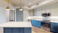 This thoughtfully designed single-floor residence combines modern aesthetics with functional living. The kitchen serves as the centerpiece of the home, showcasing a striking two-tone cabinet scheme pairing bold blue lower cabinets with light gray upper cabinets, all topped with crisp white quartz countertops. A central island with a white countertop provides additional workspace and casual dining. Stainless steel appliances — including a side-by-side refrigerator, slide-in range, built-in microwave, and dishwasher — complement the clean, contemporary look. Under-cabinet lighting and a white tile backsplash add a polished finish. A brass pendant light overhead adds a warm, stylish accent.

The bedrooms feature light wood-look flooring, neutral walls, and ceiling fans, with windows that invite natural light and views of the surrounding area. The panel-style interior doors throughout add a cohesive architectural detail.

The bathroom is a standout space, featuring rich terracotta hexagonal floor tiles contrasted against a blue vanity with white countertop and a framed mirror. A soaking tub enclosed in classic white subway tile completes the bath. An adjacent walk-in closet with built-in shelving and hanging rods offers generous storage. Light wood-look flooring flows throughout the living areas, tying the entire space together with warmth and continuity.