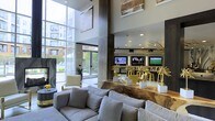 This single-floor space presents a striking contemporary lounge environment defined by its dramatic double-height ceilings and expansive floor-to-ceiling windows that flood the interior with natural light. The glazed facade offers views of a landscaped outdoor area, creating a seamless visual connection between inside and out. The seating area is anchored by a generously sized sectional sofa in neutral grey upholstery, complemented by accent chairs in soft tones, establishing a welcoming and comfortable gathering space. A standout feature is the live-edge wood counter or display surface, which adds an organic, artisanal contrast to the otherwise sleek, modern aesthetic. Atop it sit bold gold sculptural decorative pieces that serve as eye-catching focal points. Recessed lighting and multiple wall-mounted flat-screen televisions are integrated into a dark feature wall, enhancing both functionality and visual interest. Large-scale artwork adorns the upper walls, contributing to the gallery-like atmosphere. The overall ambiance balances sophistication with comfort, combining natural materials, curated décor, and modern architecture into a cohesive and inviting communal environment.