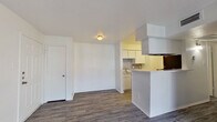 This single-floor, one-bedroom, one-bathroom unit offers a practical and efficiently designed layout across a modest footprint. The combined kitchen and living area anchors the main space, featuring durable hardwood-style flooring in a cool gray tone that runs throughout. The kitchen is defined by a peninsula-style breakfast bar, white cabinetry, a tile backsplash with a subtle geometric pattern, and black appliances including a full-size refrigerator. A dedicated dining area sits adjacent, providing a natural transition between the kitchen and living spaces. The living room benefits from a sliding glass door fitted with vertical blinds, allowing for natural light and outdoor access. A separate dining area connects smoothly to the main living space. The bedroom is generously proportioned, featuring plush carpeting, a large window with horizontal blinds that brings in natural light, and a closet with its own door. The bathroom is clean and functional, equipped with a full-size soaking tub and shower combination with white tile surround. Throughout the unit, white walls and textured ceilings create a bright, neutral backdrop. A hallway provides connectivity between the private and shared areas of the home.