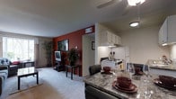 This single-floor residence showcases an open-concept layout that seamlessly connects the dining, living, and kitchen areas into one cohesive living environment. The dining area is anchored by a granite-style table surrounded by dark leather chairs, set with decorative red bowls and glassware, creating an inviting atmosphere for meals and entertaining. Adjacent to the dining space, the kitchen features white cabinetry, a gas range with an overhead range hood, and a full-size refrigerator, offering functional cooking space within easy reach of the dining area. A ceiling fan overhead provides both lighting and air circulation throughout the open space. The living area, visible beyond a doorway, is carpeted and features a bold deep-red accent wall that adds a striking visual contrast to the neutral tones of the rest of the space. A flat-screen television sits on a compact media stand, complemented by decorative indoor plants that bring natural elements into the room. The overall ambiance is warm and practical, combining everyday functionality with thoughtful design details throughout.