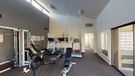 This single-floor fitness center, located in Los Angeles, CA 91343, is a well-equipped community amenity space designed to support a range of workout routines. The facility features impressively tall ceilings that create an open, airy atmosphere, complemented by a row of clerestory windows positioned near the roofline that allow natural light to filter into the space throughout the day.

The main workout area is outfitted with multiple treadmills, elliptical machines, a rowing machine, a multi-station cable and strength training unit branded Pacific, and a flat utility bench, providing users with both cardio and resistance training options. The flooring throughout the exercise area is a durable, textured carpet suitable for gym use, and the walls are finished in a neutral two-tone color scheme of warm beige and charcoal gray.

A large mirror along one wall enhances the sense of space while allowing users to monitor their form. A sliding glass door provides direct access to an outdoor area with visible greenery, bringing a connection to the outdoors into the workout environment. The space also includes climate control via ceiling-mounted ventilation units.

Adjacent to the main fitness area is a carpeted hallway corridor that leads to additional rooms, as well as a glassed-in mailroom equipped with individual resident mailboxes, reflecting this facility's role as part of a larger residential community.