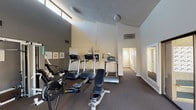 This single-floor fitness center, located in Los Angeles, CA 91343, is a well-equipped community amenity space designed to support a range of workout routines. The facility features impressively tall ceilings that create an open, airy atmosphere, complemented by a row of clerestory windows positioned near the roofline that allow natural light to filter into the space throughout the day.

The main workout area is outfitted with multiple treadmills, elliptical machines, a rowing machine, a multi-station cable and strength training unit branded Pacific, and a flat utility bench, providing users with both cardio and resistance training options. The flooring throughout the exercise area is a durable, textured carpet suitable for gym use, and the walls are finished in a neutral two-tone color scheme of warm beige and charcoal gray.

A large mirror along one wall enhances the sense of space while allowing users to monitor their form. A sliding glass door provides direct access to an outdoor area with visible greenery, bringing a connection to the outdoors into the workout environment. The space also includes climate control via ceiling-mounted ventilation units.

Adjacent to the main fitness area is a carpeted hallway corridor that leads to additional rooms, as well as a glassed-in mailroom equipped with individual resident mailboxes, reflecting this facility's role as part of a larger residential community.