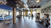 The fitness center at The Lofts at Helmetta is a well-appointed, single-floor facility that blends industrial design with a fully equipped workout environment. The space is defined by its exposed ceiling, featuring large silver ductwork, open beams, and track lighting that collectively contribute to a modern loft aesthetic. Durable rubber flooring spans the workout areas, providing a practical and slip-resistant surface throughout.

The cardio section is generously stocked with True-brand treadmills, ellipticals, and stationary bikes — including upright and recumbent models — arranged to allow comfortable spacing between users. Large windows along the perimeter invite natural light and offer outdoor views, enhancing the overall atmosphere. A ceiling fan provides additional air circulation in the cardio zone.

The strength training area includes a multi-station cable machine, adjustable weight bench, a full rack of dumbbells, and stability balls, catering to a variety of workout routines. Floor-to-ceiling mirrors along one wall expand the sense of space and assist with form checks during exercise. Mounted flat-screen televisions are positioned for convenient viewing throughout the room.

A decorative bicycle wheel wall installation adds a creative, thematic touch that ties into the loft's character. A water fountain is conveniently located near the entrance, completing this comprehensive and thoughtfully designed fitness facility.
