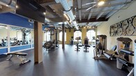 The fitness center at The Lofts at Helmetta is a well-appointed, single-floor facility that blends industrial design with a fully equipped workout environment. The space is defined by its exposed ceiling, featuring large silver ductwork, open beams, and track lighting that collectively contribute to a modern loft aesthetic. Durable rubber flooring spans the workout areas, providing a practical and slip-resistant surface throughout.

The cardio section is generously stocked with True-brand treadmills, ellipticals, and stationary bikes — including upright and recumbent models — arranged to allow comfortable spacing between users. Large windows along the perimeter invite natural light and offer outdoor views, enhancing the overall atmosphere. A ceiling fan provides additional air circulation in the cardio zone.

The strength training area includes a multi-station cable machine, adjustable weight bench, a full rack of dumbbells, and stability balls, catering to a variety of workout routines. Floor-to-ceiling mirrors along one wall expand the sense of space and assist with form checks during exercise. Mounted flat-screen televisions are positioned for convenient viewing throughout the room.

A decorative bicycle wheel wall installation adds a creative, thematic touch that ties into the loft's character. A water fountain is conveniently located near the entrance, completing this comprehensive and thoughtfully designed fitness facility.