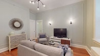 This single-floor unit blends classic architectural character with contemporary furnishings throughout its thoughtfully arranged layout. The living room serves as the centerpiece, showcasing notably tall ceilings, soft sage-green panel-detailed walls, and warm hardwood flooring that runs throughout the main spaces. A striking multi-arm sputnik-style chandelier anchors the ceiling, complemented by sleek wall sconces that provide layered ambient lighting. A sectional sofa, accent chair, and a striped coffee table create a comfortable, well-organized seating area, while a window fitted with blinds allows natural light to fill the room.

The bedroom continues the calm, muted color palette with light blue-gray walls and hardwood floors. A double window admits generous natural light and houses a window air conditioning unit, while a cast-iron radiator speaks to the unit's classic construction. The bedroom offers comfortable accommodations with ample space for furniture.

The kitchen features warm honey-toned wood cabinetry with granite-effect countertops, a gas range, and a stainless steel sink—providing practical functionality. The bathroom is finished in clean white tile wainscoting, with a tub-shower combination, pedestal sink, and a bright window above a traditional cast-iron radiator, reinforcing the timeless character present throughout the space.