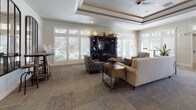 This expansive single-floor clubhouse is thoughtfully designed to offer a warm and inviting atmosphere across a generous open floor plan. The centerpiece of the main lounge is a striking floor-to-ceiling stacked stone fireplace, flanked by multiple seating arrangements that encourage relaxed gathering. Tray ceilings with decorative wood trim add architectural elegance, while ceiling fans and recessed lighting ensure the space remains comfortable and well-lit.

Throughout the space, wood-look flooring transitions seamlessly between areas, complemented by textured area rugs that define individual seating zones. Large windows and glass-paned French doors line the walls, flooding the interior with natural light and offering views of the lush outdoor surroundings. A second lounge area features a built-in entertainment center and additional seating, making it ideal for casual use.

The kitchen is well-appointed with rich dark cabinetry, granite countertops, a tile backsplash, and under-cabinet lighting. A breakfast bar with upholstered bar seating and elegant pendant lighting provides a welcoming spot for casual dining. The dining area is anchored by a solid wood table beneath a decorative chandelier, with oversized windows bathing the room in natural light. Overall, the space exudes a refined yet comfortable ambiance suited for community events and everyday gatherings alike.