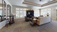 This expansive single-floor clubhouse is thoughtfully designed to offer a warm and inviting atmosphere across a generous open floor plan. The centerpiece of the main lounge is a striking floor-to-ceiling stacked stone fireplace, flanked by multiple seating arrangements that encourage relaxed gathering. Tray ceilings with decorative wood trim add architectural elegance, while ceiling fans and recessed lighting ensure the space remains comfortable and well-lit.

Throughout the space, wood-look flooring transitions seamlessly between areas, complemented by textured area rugs that define individual seating zones. Large windows and glass-paned French doors line the walls, flooding the interior with natural light and offering views of the lush outdoor surroundings. A second lounge area features a built-in entertainment center and additional seating, making it ideal for casual use.

The kitchen is well-appointed with rich dark cabinetry, granite countertops, a tile backsplash, and under-cabinet lighting. A breakfast bar with upholstered bar seating and elegant pendant lighting provides a welcoming spot for casual dining. The dining area is anchored by a solid wood table beneath a decorative chandelier, with oversized windows bathing the room in natural light. Overall, the space exudes a refined yet comfortable ambiance suited for community events and everyday gatherings alike.