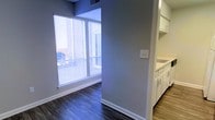 This contemporary two-floor space is thoughtfully designed with a clean, modern aesthetic that balances functionality and style. Dark hardwood flooring runs throughout, providing a rich contrast against the light gray walls and crisp white trim. Large floor-to-ceiling windows flood the interior with abundant natural light while framing urban city views, creating an open and airy atmosphere that connects the indoors with the surrounding cityscape. Horizontal blinds on the windows offer adjustable light control and privacy. The kitchen area features white cabinetry with sleek hardware, offering generous storage and a bright, organized workspace. Built-in open shelving adds both practicality and a design element to the kitchen zone. Neutral gray walls are carried consistently throughout the space, lending a cohesive and modern feel to every room. The two-floor layout provides a natural separation of living areas, allowing for distinct functional zones across each level. Ceiling vents are thoughtfully integrated to maintain a clean architectural look. The overall ambiance is polished and understated, making this a versatile space well-suited for a variety of uses.