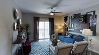 This single-floor living space showcases a thoughtfully designed interior that blends modern aesthetics with eclectic personality. The main living area is anchored by a bold blue sofa adorned with patterned throw pillows, complemented by a white accent chair and a geometric-patterned area rug in blue and white tones that adds visual depth to the room.

A ceiling fan with an integrated light fixture is centrally mounted, providing both function and a finished look to the space. Natural light enters generously through a large sliding glass door fitted with dark curtain panels, which opens to a private balcony and offers views of the surrounding area.

The accent wall features an artistic arrangement of decorative lettering, mirrors, and mixed-media artwork, giving the room a curated, gallery-like feel. Warm ambient lighting from a floor lamp adds a welcoming glow to the space during evening hours.

Lush indoor plants are positioned near the entrance area, introducing a natural element that softens the contemporary design. Dark hardwood-style flooring grounds the space and provides a rich contrast to the lighter furnishings and walls, creating a cohesive and inviting atmosphere throughout.