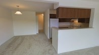 This single-floor residential unit offers a practical and cohesive open-concept layout designed for comfortable everyday living. The kitchen is positioned at the heart of the space, featuring a peninsula-style counter that provides both a food preparation surface and a natural divider between the kitchen and the adjoining living area. Wood-toned upper cabinetry offers ample storage, while white appliances, including a refrigerator and a cooktop with an overhead range hood, provide essential functionality. The countertops are finished in a neutral tone that complements the overall color palette of the space. The living area extends seamlessly from the kitchen, finished with wall-to-wall carpeting in a light beige tone that adds warmth underfoot and contributes to a soft, understated ambiance throughout. Neutral white walls and low-profile ceilings keep the interior feeling clean and uniform. A central hallway connects the main living and kitchen area to additional rooms beyond, suggesting a thoughtfully organized floor plan. Ceiling-mounted lighting provides consistent illumination throughout. Overall, the space presents a straightforward, well-maintained interior with a neutral palette suited to a variety of personal styles.