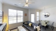 This single-floor residence offers a comfortable and well-appointed living experience with a thoughtful, open layout. The living area serves as the heart of the home, bathed in natural light streaming through multiple large windows dressed with horizontal blinds. A ceiling fan with integrated lighting hangs overhead, adding both practicality and a clean, classic touch to the space. Neutral-toned walls provide a versatile backdrop that complements the warm hardwood-style flooring below. The living room is anchored by a sleek tufted leather sofa accented with yellow and gray throw pillows, creating an inviting atmosphere with a contemporary flair. A patterned area rug in coordinating tones ties the seating area together, while a cushioned ottoman adds functional style. Adjacent to the living space, a dining area is visible, offering a seamless transition for entertaining and everyday living. A doorway with a full-light glass door connects the interior to the outside, further enhancing the sense of openness and allowing additional natural light to filter in. Crown molding details along the ceiling add an architectural refinement throughout, elevating the overall character of this bright and stylish residence.
