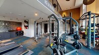This well-appointed two-level fitness center offers a comprehensive workout experience spread across two thoughtfully designed floors. The lower level serves as the primary strength and cardio zone, featuring an impressive array of equipment including squat racks, cable machines, weight benches, treadmills, ellipticals, and stationary bikes. Large floor-to-ceiling windows flood this level with abundant natural light while framing views of the surrounding greenery, creating an energizing atmosphere. The space is finished with a vibrant patchwork carpet in shades of teal, gray, and earth tones, adding a dynamic visual energy to the environment. Decorative laser-cut wall panels and bold motivational signage — including the phrase "Reach New Heights" — reinforce the facility's contemporary aesthetic. An open-railing staircase connects the two levels, maintaining a sense of visual continuity throughout. The upper level provides additional workout space, complete with medicine ball storage, benches, cardio machines, and stretching areas. Unique woven pendant light fixtures and track lighting add warmth and style. The upper floor overlooks the lower level through open railings, enhancing the spacious, double-height feel of the facility as a whole.