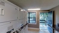 This shared laundry facility is designed to accommodate multiple users efficiently, offering a well-organized layout with a row of commercial-grade, top-loading washing machines and a bank of stacked dryers along the opposing wall. The machines are coin-operated, providing straightforward access for residents. Instructional signage is posted prominently on the walls, offering washing guidance in multiple languages for user convenience. A dark-surfaced folding counter runs along one wall, providing a practical surface for sorting and folding laundry. The room is finished with neutral taupe-toned walls, light-colored flooring, and a textured ceiling with fluorescent lighting that ensures the space is well-lit. A standout feature is the generous natural light that enters through large, black-framed windows and a glass door that opens to an outdoor deck or walkway, offering pleasant views of surrounding mature trees and greenery. A ventilation unit on the wall supports air circulation throughout the space. The overall atmosphere is functional and practical, with a clean, maintained appearance suited for shared residential use.