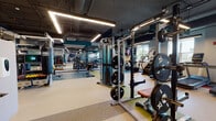 Located at 87 Union St in New Haven, CT, this single-floor fitness center is a well-appointed, contemporary gym designed to accommodate a wide range of training needs. The space is organized into distinct functional zones, each thoughtfully laid out to maximize usability and flow.

The strength training area is stocked with a comprehensive rack of dumbbells, multiple adjustable benches, and a full set of plate-loaded barbell stations with squat racks. Floor-to-ceiling mirrors line the walls, enhancing the sense of openness throughout. The cardio zone features a row of Matrix treadmills and elliptical machines positioned near large windows that bring in natural light and outdoor views, creating an energizing atmosphere for endurance training.

A dedicated functional fitness area offers TRX suspension trainers, plyo boxes, battle ropes, and a heavy punching bag, supported by colorful, durable rubber flooring in bold geometric patterns of green, orange, blue, and tan. An enclosed studio space with glass walls is available for group classes or personal training sessions.

The industrial-style ceiling exposes ductwork and structural beams painted in dark teal and black, complemented by bright linear LED lighting. Motivational large-scale wall murals — including a striking black-and-white portrait with an inspirational quote — add character and energy to the environment. The overall ambiance is dynamic, modern, and purpose-built for serious fitness.