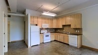 This single-floor space showcases an open-concept kitchen that serves as a central feature of the layout. The kitchen is outfitted with light maple wood cabinetry extending to the ceiling, providing generous storage capacity across both upper and lower cabinets. Dark laminate countertops contrast cleanly against the lighter wood tones, while a tile backsplash adds a subtle textural element along the walls. White appliances — including a refrigerator, electric range, built-in microwave, and dishwasher — maintain a cohesive, clean look throughout the space. The polished concrete flooring runs throughout, lending an industrial character that complements the utilitarian aesthetic of the interior. Overhead, industrial-style track lighting and a fluorescent fixture illuminate the kitchen area effectively. The ceiling height adds a sense of openness and volume to the space. A partially visible hallway or entryway at the rear of the kitchen suggests a functional layout with additional rooms beyond. Overall, the space combines practical design with understated industrial elements, creating a functional and straightforward living environment.