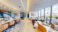 Located on Biscayne Boulevard in Miami, FL, this contemporary single-floor amenity and coworking space offers a well-appointed environment designed for both productivity and comfort. The layout spans a generous open floor plan that seamlessly integrates multiple functional zones, including a large conference area, lounge seating, and a series of glass-enclosed private meeting rooms.

The conference area anchors one side of the space and features a substantial light wood communal table surrounded by tan leather rolling chairs, set beneath stylish globe pendant lighting. A bold, colorful abstract mural serves as a dramatic focal point on the accent wall, complemented by a wall-mounted display screen. Adjacent to the conference zone, a relaxed lounge area is furnished with contemporary curved seating in warm orange and neutral tones, arranged around low coffee and console tables atop layered area rugs.

A corridor of glass-walled private offices and meeting rooms lines one side, offering privacy without sacrificing visual openness. These rooms feature dark-framed glass partitions, patterned carpet, and individual display screens. Light wood flooring runs throughout the open areas, reflecting the abundant natural light that pours in through floor-to-ceiling windows along the exterior wall. These expansive windows frame open outdoor views, enhancing the airy, sun-filled ambiance characteristic of Miami's vibrant atmosphere. Recessed and cove lighting add a polished, professional finish to the overall design.