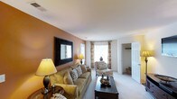 This single-floor residence offers a thoughtfully designed and welcoming interior that balances comfort with style. The living room serves as the heart of the space, showcasing warm earth-toned accent walls that create a cozy and inviting atmosphere. Plush wall-to-wall carpeting runs throughout, adding softness underfoot and contributing to the overall sense of warmth. Large windows dressed with elegant floral-patterned drapes allow generous natural light to fill the room, enhancing its open and airy feel. The space is well-suited for relaxed living, with ample room for a full-sized sofa, accent chairs, and a central coffee table. A decorative framed mirror mounted on the accent wall adds depth and visual interest to the room. Recessed lighting in the ceiling complements the natural light, ensuring the space remains bright at any hour. A doorway visible from the living area leads to what appears to be an additional room or walk-in closet, hinting at further functional spaces within the layout. Overall, the residence presents a harmonious blend of neutral tones, natural light, and thoughtful design elements that make it both practical and aesthetically pleasing.