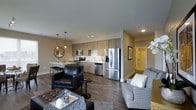 This single-floor residence showcases a thoughtfully designed open-concept layout that integrates the living, dining, and kitchen areas into one cohesive, flowing space. The kitchen is outfitted with stainless steel appliances, including a French door refrigerator, dishwasher, and microwave, all complemented by flat-front cabinetry in a warm light wood finish. A clean white countertop with an undermount sink adds a polished touch, while track lighting and pendant lights overhead provide focused illumination throughout the space. The dining area features a sleek bar-height counter with seating, ideal for casual meals. The living area is generously proportioned, accommodating a mix of seating styles, including a leather accent chair and a patterned sofa, arranged over a soft area rug. Dark hardwood-style flooring runs throughout, grounding the neutral beige walls and grey ceiling. A hallway visible in the background leads to additional areas, including what appears to be a well-lit bathroom. The overall ambiance is modern, clean, and functional, with a warm and inviting character.
