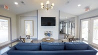 Located at 6520 Dorchester Rd in North Charleston, South Carolina, this single-floor space showcases a thoughtfully curated interior with a welcoming, coastal-modern aesthetic. The main living area immediately draws attention with its soaring vaulted ceiling, which lends an open and airy quality to the room. A striking shiplap accent wall serves as the central focal point, framing both a built-in fireplace and a wall-mounted flat-screen television, blending comfort and style seamlessly. A mid-century modern chandelier with brass arms and globe bulbs adds a warm, refined touch overhead. Natural light flows generously through the space, highlighting the smooth hardwood-style flooring that runs throughout. The living area is furnished with a deep navy blue sectional sofa, a rustic round wooden coffee table, and a pair of classic striped armchairs, creating an inviting and relaxed atmosphere. Tasteful coastal artwork, including a framed lighthouse print in gold tones, reinforces the nautical design theme. A large mirror along one wall amplifies the sense of space and reflects additional natural light. Sliding glass doors visible in the background suggest seamless access to an outdoor area, further connecting the interior to its surroundings. Overall, the space offers a polished yet comfortable environment with strong architectural character.