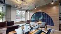 This two-floor space masterfully blends industrial architecture with vibrant, artistic design. The ground floor opens with polished concrete floors and soaring exposed ceilings that reveal structural ductwork and piping, lending a raw yet refined aesthetic. A dramatic large-scale bird mural set against a teal ombre backdrop anchors the entry corridor, complemented by sculptural branch installations that extend across the ceiling. Glass-partitioned private offices line one side, maintaining visual openness throughout.

The main lounge area is a standout, featuring a striking arched fireplace clad in deep cobalt blue tile set within a warm wood surround. A sculptural multi-arm chandelier hangs overhead, illuminating a bold geometric patterned area rug and mid-century modern seating. Adjacent, a generous dining and gathering space includes a live-edge wood communal table, blue tile wet bar with open wood shelving, and floor-to-ceiling windows that flood the room with natural light and outdoor views.

The second floor offers additional lounge and collaborative seating areas finished in rich teal with curated artwork, overlooking the ground floor via an industrial mesh-rail mezzanine. An upper-level dining room with abundant natural light, patterned carpet, and varied seating configurations completes this thoughtfully designed, multifunctional space.