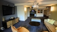 This single-floor amenity lounge is thoughtfully designed to serve as a vibrant social and recreational hub. The main seating area sets a contemporary tone with plush tan leather chairs, a sleek round black coffee table, and a padded bench ottoman arranged around a wall-mounted flat-screen TV. The space is anchored by a striking sculptural chandelier with an abstract, multi-pronged metallic design that serves as a bold focal point overhead. Warm wall sconces and recessed lighting complement the overall ambiance, casting an inviting glow throughout. Patterned low-pile carpeting in deep blue-grey tones runs underfoot, adding texture and visual depth to the interior. A rustic wood-and-metal credenza adds an industrial-chic accent along one wall, while a pair of framed artworks in rich tones bring color and character to the room. Beyond the main lounge, an open passage leads to a dedicated game room featuring a full-size billiards table and an additional wall-mounted television, making it an ideal space for entertainment. The open layout and connected zones create a seamless flow, making the overall space well-suited for casual gatherings, leisure activities, and community engagement.