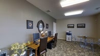 The Glenmary Village business center offers a well-appointed, single-floor space designed to meet the everyday professional needs of its residents. The room features a warm, neutral color palette with soft gray walls that create a calm and focused atmosphere throughout.

At the heart of the space sits a rich wooden desk accommodating dual computer workstations, paired with elegant high-back leather chairs with nailhead trim detailing. The desk is complemented by a warm brass desk lamp, adding a refined touch to the functional setup. A multifunction copier and printer station is conveniently positioned along the far wall, ensuring residents have easy access to essential office equipment.

The space is finished with patterned carpeting in earthy tones that adds visual interest underfoot, while vaulted ceilings contribute to an open, airy feel. Overhead fluorescent lighting provides ample illumination throughout the room. Decorative framed artwork with an equestrian theme adorns the walls, adding character and a cohesive design aesthetic. An oval decorative mirror serves as a focal accent piece, enhancing the room's charm.

Overall, the Glenmary Village business center balances practicality with a welcoming, tastefully decorated environment ideal for work and productivity.