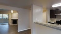 This single-story space features an open, flowing layout that connects the kitchen and living areas seamlessly. A half-wall partition with a granite countertop serves as a breakfast bar, providing a natural boundary between the kitchen and the adjoining spaces while maintaining an open feel. The neutral-toned walls throughout create a clean, versatile backdrop. Wood-look flooring extends through the main areas, adding warmth and visual continuity. The living area, visible through a wide doorway, features a fireplace that serves as a focal point. Recessed outlets and light switches are positioned throughout, reflecting a practical and functional design. The overall layout benefits from interconnected rooms that allow for easy movement and a cohesive atmosphere throughout the space.