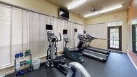 Located at 2704 Salem Church Rd in Fredericksburg, Virginia, this single-floor fitness center offers a well-appointed space designed to support cardio-focused workouts. The room features a suite of modern exercise equipment, including multiple treadmills, an elliptical trainer, a recumbent bike, and an upright stationary bike, each equipped with its own personal display monitor for an enhanced workout experience. A wall-mounted flat-screen television provides additional entertainment options for users during their sessions. The space is illuminated by a combination of overhead fluorescent lighting and natural light streaming through several windows fitted with white horizontal blinds, creating a bright and energizing atmosphere. A ceiling fan adds to the comfort of the room during use. Durable rubber flooring covers the entire workout area, providing both safety and stability. Elegant French doors with transom windows connect the fitness room to an adjoining lounge or common area, adding an architectural detail that enhances the overall character of the space. A posted fitness rules sign and modest greenery contribute to the room's functional yet welcoming environment.