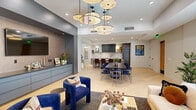 Located at 8555 East Raintree Drive in Scottsdale, Arizona, this thoughtfully designed single-floor space seamlessly integrates a fully equipped kitchen and an expansive living area into one cohesive, open-plan layout. Light hardwood flooring flows throughout, unifying the space and enhancing its bright, airy character.

The kitchen is a standout feature, anchored by a dramatic marble island with waterfall edges and seating for multiple guests on rich burgundy leather bar stools. Dark, fluted cabinetry lines the walls, complemented by a full marble backsplash, integrated appliances, and open shelving. A large dining table with sleek navy-and-gold chairs occupies the central zone, bridging the kitchen and living areas.

Overhead, a striking sculptural chandelier composed of multiple disc-shaped pendants serves as the visual centerpiece of the space, adding an artistic, avant-garde quality to the interior.

The living area is defined by a generous cream sofa, a burl wood coffee table, and deep blue accent chairs. A built-in credenza with a wall-mounted display screen anchors one side of the room. Floor-to-ceiling sheer drapery lines an entire wall of glazing, filtering the abundant Arizona sunlight while offering a refined, hotel-inspired ambiance throughout.