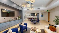 Located at 8555 East Raintree Drive in Scottsdale, Arizona, this thoughtfully designed single-floor space seamlessly integrates a fully equipped kitchen and an expansive living area into one cohesive, open-plan layout. Light hardwood flooring flows throughout, unifying the space and enhancing its bright, airy character.

The kitchen is a standout feature, anchored by a dramatic marble island with waterfall edges and seating for multiple guests on rich burgundy leather bar stools. Dark, fluted cabinetry lines the walls, complemented by a full marble backsplash, integrated appliances, and open shelving. A large dining table with sleek navy-and-gold chairs occupies the central zone, bridging the kitchen and living areas.

Overhead, a striking sculptural chandelier composed of multiple disc-shaped pendants serves as the visual centerpiece of the space, adding an artistic, avant-garde quality to the interior.

The living area is defined by a generous cream sofa, a burl wood coffee table, and deep blue accent chairs. A built-in credenza with a wall-mounted display screen anchors one side of the room. Floor-to-ceiling sheer drapery lines an entire wall of glazing, filtering the abundant Arizona sunlight while offering a refined, hotel-inspired ambiance throughout.