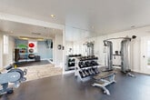 Located at 575 McDonough Pkwy in McDonough, Georgia, this fitness center offers a thoughtfully designed, single-floor layout divided into distinct functional zones. The space features a clean, modern aesthetic with neutral-toned walls, recessed lighting, and a combination of dark rubber flooring in the equipment areas and herringbone-patterned carpet tile in the transitional zones.

The cardio section is a standout area, lined with an impressive wall of French doors and windows that flood the room with natural light and offer pleasant outdoor views. Treadmills, an elliptical, and a stationary bike are positioned to take full advantage of this bright, airy atmosphere, accompanied by wall-mounted televisions for entertainment.

The strength training zone features a large floor-to-ceiling mirror that visually expands the space, alongside a multi-function cable machine, a weight bench, and a well-stocked dumbbell rack. A dedicated flexibility and functional fitness area provides carpeted flooring and a rack of stability balls and medicine balls, ideal for stretching and dynamic workouts.

A central transition corridor connects the zones and includes a water fountain station set against a decorative geometric accent wall. The fitness center also benefits from direct exterior access via French-style glass doors, providing excellent ventilation and convenience for users.