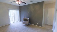 This single-story space features a well-appointed living area that combines comfort and style. The room is anchored by a striking gray accent wall, which creates a bold focal point against the lighter surrounding walls and white baseboards. Soft, neutral-toned carpeting extends throughout, adding warmth underfoot and contributing to the room's comfortable ambiance.

A ceiling fan with a built-in light fixture is centrally positioned overhead, providing both air circulation and supplemental lighting. Natural light enters generously through a large sliding glass door fitted with vertical blinds, offering flexibility in light control and access to what appears to be an outdoor area or balcony beyond.

The layout provides an open, airy feel with ample wall space suitable for a variety of furniture arrangements. The textured ceiling adds a subtle decorative element, while the overall neutral color palette creates a versatile backdrop adaptable to various design styles. The combination of natural light, neutral tones, and the distinctive accent wall makes this living space both functional and visually appealing.