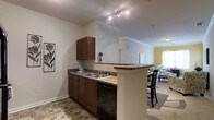 This single-floor residence offers an open-concept design that seamlessly connects the kitchen, dining, and living areas, creating a cohesive and functional living environment. The kitchen is a standout feature, equipped with rich dark wood cabinetry that provides ample storage, stone-look countertops, a double-basin sink, and a black dishwasher. A peninsula with a breakfast bar extends into the dining area, making it ideal for casual dining or entertaining. Track lighting above the kitchen adds focused illumination to the workspace. The flooring in the kitchen features a slate-style tile, adding texture and visual interest to the space. Transitioning into the dining and living areas, the flooring shifts to plush carpeting, creating a warm and inviting atmosphere. Crown molding accents the ceilings throughout, lending a refined architectural detail to the interior. The living area benefits from natural light filtering in through the windows, complemented by neutral wall tones that provide a versatile backdrop. The open sightlines between rooms enhance the sense of space, making the overall layout feel expansive and well-connected throughout.