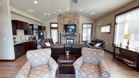 Located at 1605 North River Ridge Boulevard in Spokane, WA, this single-floor community space is thoughtfully designed to offer comfort and functionality in an inviting atmosphere. The centerpiece of the main gathering area is a striking floor-to-ceiling stacked stone fireplace, complete with a wood mantel and wall-mounted flat-screen television, anchoring the room with warmth and visual impact. Soaring vaulted ceilings with recessed lighting amplify the sense of openness, while hardwood-style flooring runs throughout, lending a cohesive and polished look. Generous seating, including leather reclining sofas and upholstered accent chairs, is arranged to encourage conversation and relaxation. Large windows on multiple walls invite abundant natural light and provide views of the surrounding exterior. A dining area with a wood table and chairs sits adjacent to the main lounge, ideal for group gatherings. The kitchen is well-appointed with rich dark cabinetry, granite-look countertops, stainless steel appliances including a refrigerator, range, and microwave, and a double sink. A water fountain and ADA-accessible restroom are also conveniently located within the space, reflecting attention to accessibility and guest comfort throughout.