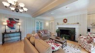 This single-floor clubhouse offers a warm and inviting atmosphere designed for community gathering and relaxation. The main lounge area is anchored by a classic wood-burning fireplace with a white mantel, seasonally decorated and flanked by lush potted greenery. Vertical shiplap-style wall paneling in soft white tones lends a charming, cottage-inspired character throughout, while exposed ceiling beams add architectural interest and depth. An elegant chandelier illuminates the center of the room, complementing wall sconces that cast a soft, welcoming glow. Light hardwood flooring runs throughout the space, contributing to the bright and airy feel. Comfortable upholstered sofas and a floral area rug define the seating area, creating an intimate space ideal for socializing. Multiple sets of French doors open to views of the surrounding greenery, filling the interior with natural light and providing easy access to the outdoors. A separate dining area features a round wood table with ladder-back chairs, positioned near its own set of French doors for additional natural light. A wrought iron railing detail and an open lower-level landing add a subtle architectural accent to the space. Overall, the clubhouse exudes a relaxed, residential charm suited for community events and casual gatherings alike.