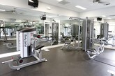 This fitness center is a single-level facility designed to support a comprehensive workout experience. The space is outfitted with a range of strength training equipment, including cable machines, multi-function weight stations, and bench press setups from brands such as Body-Solid and Matrix. Rubber flooring covers the workout area, providing both durability and safety underfoot.

Large mirrors line multiple walls throughout the space, visually expanding the room and allowing users to monitor their form during exercise. Ceiling fans are mounted overhead, complementing the flush-mounted fluorescent lighting to maintain a comfortable environment. Wall-mounted flat-screen televisions are positioned for easy viewing during workouts.

Natural light enters through generously sized windows and glass-paneled double doors, which appear to open toward an outdoor pool area, creating a bright and open atmosphere. The walls are finished in a clean, neutral gray, and white trim detailing adds a polished, classic touch to the overall aesthetic. The layout is open and thoughtfully arranged, allowing users to move between equipment with ease. An adjacent room provides additional equipment space, accessible through a framed interior doorway, further enhancing the overall capacity and versatility of the fitness center.