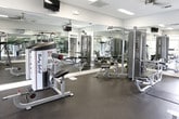 This fitness center is a single-level facility designed to support a comprehensive workout experience. The space is outfitted with a range of strength training equipment, including cable machines, multi-function weight stations, and bench press setups from brands such as Body-Solid and Matrix. Rubber flooring covers the workout area, providing both durability and safety underfoot.

Large mirrors line multiple walls throughout the space, visually expanding the room and allowing users to monitor their form during exercise. Ceiling fans are mounted overhead, complementing the flush-mounted fluorescent lighting to maintain a comfortable environment. Wall-mounted flat-screen televisions are positioned for easy viewing during workouts.

Natural light enters through generously sized windows and glass-paneled double doors, which appear to open toward an outdoor pool area, creating a bright and open atmosphere. The walls are finished in a clean, neutral gray, and white trim detailing adds a polished, classic touch to the overall aesthetic. The layout is open and thoughtfully arranged, allowing users to move between equipment with ease. An adjacent room provides additional equipment space, accessible through a framed interior doorway, further enhancing the overall capacity and versatility of the fitness center.