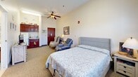 Located at 3980 S Jackson Dr in Independence, MO, this single-bedroom suite offers a well-organized and comfortable living environment within a residential care setting. The main bedroom is generously proportioned, featuring soft carpeted flooring, neutral-toned walls, and a ceiling fan that contribute to a calm and welcoming ambiance. A large window with a decorative valance and blinds allows natural light to fill the room while providing outdoor views of the surrounding grounds. The room accommodates a full-sized bed, a reclining chair, an upholstered armchair, a multi-drawer dresser, and a flat-screen television, demonstrating thoughtful use of the available space. A small kitchenette area with wood cabinetry and a sink is accessible within the room, adding a practical convenience for residents. The bathroom is clean and functional, featuring a curtained shower area, toilet, vanity with wood cabinetry, and safety-conscious design elements including a wall-mounted pull cord and grab bar. Bright, even lighting throughout the suite ensures visibility and ease of navigation. The overall layout is designed to support daily living needs while maintaining a warm, home-like atmosphere.