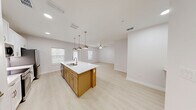 This thoughtfully designed single-story residence offers a bright, open-concept living space finished with light luxury vinyl plank flooring throughout. The kitchen serves as the heart of the home, anchored by a warm wood-toned island with a white quartz countertop, dual undermount sink, and stylish pendant lighting. White shaker cabinetry reaches toward the ceiling, complemented by stainless steel appliances including a range, microwave, and refrigerator. The kitchen flows seamlessly into the living area, where a ceiling fan adds comfort and natural light streams in through multiple windows. The generously sized primary bedroom provides a calm retreat, also equipped with a ceiling fan and ample room for furnishings. The full bathroom features a lengthy white vanity with quartz countertop, a gold-accented framed mirror, and a soaking tub with shower combination finished in clean white tile. A dedicated walk-in closet with built-in shelving and hanging rods offers practical storage solutions, while an in-unit laundry room houses a full-size washer and dryer. The exterior includes a private patio entrance with a fenced yard area and views of the surrounding community grounds.