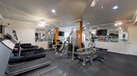 This fitness center offers a thorough and well-organized workout environment designed to accommodate a wide range of exercise routines. The space is outfitted with an extensive selection of strength-training machines, including cable systems, weight stations, and resistance equipment, providing users with the tools needed for a full-body workout. Cardio equipment, including treadmills, lines the perimeter, making efficient use of the available floor space.

The interior features a dark, rubberized flooring ideal for heavy equipment and high-traffic use. Accent walls in a bold orange hue add visual energy to the space, complementing the neutral ceiling and trim. Large wall-to-wall mirrors are installed throughout, enhancing the sense of openness and allowing users to monitor their form during exercise. Multiple ceiling fans are mounted overhead, contributing to air circulation and comfort during workouts.

Recessed lighting provides consistent, bright illumination throughout the facility. Wall-mounted flat-screen televisions offer entertainment during sessions. The single-floor layout ensures seamless accessibility, and the overall design prioritizes functionality and practicality, making this fitness center a capable and well-appointed amenity for regular use.