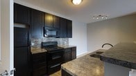 Located at 7505 Worthington Galena Rd in Columbus, Ohio, this single-floor property offers a well-appointed and thoughtfully designed living space. The kitchen is a standout feature, showcasing rich, dark espresso cabinetry that extends to the ceiling, providing generous storage throughout. Granite countertops in warm, earthy tones complement the bold cabinetry, while a coordinating tile backsplash in a linear mosaic pattern adds texture and visual interest. Black appliances, including a range and over-the-range microwave, tie the space together with a cohesive, modern aesthetic. A kitchen island or peninsula with an undermount sink and matching granite surface offers additional workspace and a natural gathering point. Track lighting overhead ensures the kitchen is well-lit and functional. Light hardwood-style flooring runs through the space, providing a warm contrast to the darker elements of the kitchen. The open layout connects the kitchen to an adjacent area, allowing for a natural flow between spaces. The overall ambiance is modern and refined, with a clean, neutral color palette that creates a welcoming and versatile interior suitable for a variety of styles and preferences.