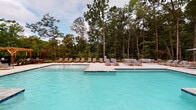 Located at 5824 McRimmon Pkwy in Morrisville, NC, this single-level community amenity space combines thoughtful architectural design with resort-inspired outdoor living. The centerpiece of the property is a generously sized outdoor swimming pool with crystal-clear blue water, set against a backdrop of towering mature trees that provide a serene, natural atmosphere. The expansive concrete sundeck surrounding the pool is well-appointed with numerous teak-style lounge chairs and seating areas, offering ample space for relaxation and socializing. A charming wooden pergola anchors one end of the pool area, adding a warm, structural accent to the outdoor landscape. The pool area is enclosed by a sleek metal fence, ensuring both safety and an unobstructed view of the surrounding woodland scenery. The interior clubhouse is accessible through prominent floor-to-ceiling glass doors framed by classic exposed brick walls, creating an inviting industrial-modern aesthetic. Sliding barn-style doors and large glass panels allow natural light to flow throughout the interior while maintaining visual connectivity to the outdoor pool deck. The combination of brick masonry, dark metal framing, and expansive glazing gives the building a distinctive, upscale character that complements the lush, wooded setting.