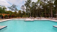 Located at 5824 McRimmon Pkwy in Morrisville, NC, this single-level community amenity space combines thoughtful architectural design with resort-inspired outdoor living. The centerpiece of the property is a generously sized outdoor swimming pool with crystal-clear blue water, set against a backdrop of towering mature trees that provide a serene, natural atmosphere. The expansive concrete sundeck surrounding the pool is well-appointed with numerous teak-style lounge chairs and seating areas, offering ample space for relaxation and socializing. A charming wooden pergola anchors one end of the pool area, adding a warm, structural accent to the outdoor landscape. The pool area is enclosed by a sleek metal fence, ensuring both safety and an unobstructed view of the surrounding woodland scenery. The interior clubhouse is accessible through prominent floor-to-ceiling glass doors framed by classic exposed brick walls, creating an inviting industrial-modern aesthetic. Sliding barn-style doors and large glass panels allow natural light to flow throughout the interior while maintaining visual connectivity to the outdoor pool deck. The combination of brick masonry, dark metal framing, and expansive glazing gives the building a distinctive, upscale character that complements the lush, wooded setting.