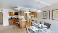 This single-floor residence offers a thoughtfully designed layout with a range of functional and comfortable spaces throughout. The kitchen is a standout feature, showcasing warm wood cabinetry, a peninsula with bar seating, and a dedicated wet bar area with a mirrored backsplash, all complemented by a decorative coffered ceiling detail that adds architectural character. The kitchen flows seamlessly into the dining area, which is bathed in natural light from a large window and anchored by a pendant light above a glass-top dining table. The living room is bright and welcoming, featuring a tile-surround fireplace as a focal point, a wall-mounted television, and sliding glass doors that open to an outdoor patio, bringing in ample natural light. The primary bedroom is generously sized with plush carpeting, direct access to an en-suite bathroom complete with a wide vanity and ample counter space, and a walk-in closet. A secondary bedroom offers a versatile space with a window, carpet flooring, and room for a desk area. The bathroom includes a full tub-shower combination. Practical conveniences include an in-unit laundry area with a full-size washer and dryer. Throughout, neutral tones and soft carpeting create a cohesive and inviting ambiance.
