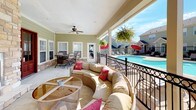 This single-level outdoor amenity space is designed for both relaxation and socializing, offering a generously sized covered patio that seamlessly connects to a resort-style community pool area. The patio is equipped with ceiling fans, providing comfortable airflow during warm days, and is enclosed by decorative wrought-iron railings that lend an elegant yet secure feel to the space.

The covered patio features multiple distinct seating zones: a large dining table with slatted chairs accommodates group gatherings, while a circular wicker sectional sofa with vibrant accent pillows creates an inviting lounge area with direct views of the sparkling outdoor pool. A separate bistro-style table and chairs provide an additional intimate seating option.

A standout architectural feature is the floor-to-ceiling natural stone fireplace, flanked by rich wood-framed French doors with glass panes that lead into the interior. The stone accents continue along the base of the exterior walls, adding warmth and texture to the overall design.

The pool area is visible from the patio and is surrounded by a concrete deck furnished with lounge chairs and red market umbrellas, set against a backdrop of lush green lawns and neighboring residential buildings. The combination of covered comfort, open-air views, and multiple gathering spots makes this a highly functional and inviting community outdoor space.