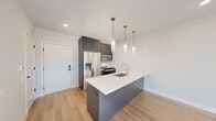 This contemporary single-floor unit showcases a thoughtfully designed open-concept layout with clean lines and modern finishes throughout. The kitchen serves as the centerpiece of the living space, anchored by a sleek peninsula island topped with a crisp white countertop and fitted with an undermount sink and a brushed gold faucet. Dark-toned cabinetry provides strong visual contrast against the white surfaces, while three cylindrical pendant lights with gold hardware hang above, adding a refined accent. Stainless steel appliances, including a French door refrigerator, microwave, range, and dishwasher, complete the kitchen's functional appeal. Light hardwood flooring runs throughout the main living areas and bedrooms, contributing warmth and continuity. Large floor-to-ceiling windows in the living and bedroom areas invite generous natural light and frame views of the surrounding urban streetscape. Each bedroom offers a clean, neutral palette with recessed lighting and a built-in closet featuring shelving and hanging storage. The bathroom is finished with hexagonal floor tiles, a floating white vanity, a soaking tub, and marble-style wall tiles accented by matte black fixtures and hardware, creating a crisp, spa-like atmosphere.