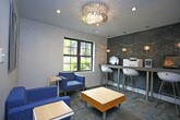 This single-floor business center is thoughtfully designed to provide a functional and visually striking workspace. The room is anchored by a long, streamlined counter-height desk finished in a clean light gray, offering ample workspace for multiple users simultaneously. The desk is equipped with desktop computers and a multifunction printer, making it a fully operational workstation for a variety of professional needs.

Overhead, a trio of crystal-style pendant lights hang at evenly spaced intervals, casting warm, ambient light across the workspace and adding a touch of elegance to the utilitarian setting. Modern white sculpted bar-height chairs are tucked neatly beneath the desk, combining comfort with contemporary style.

The defining feature of the room is the dramatic textured accent wall behind the workstation, featuring a richly layered stone or tile pattern in deep charcoal, slate blue, and silver tones. This bold backdrop adds depth and sophistication, transforming what might otherwise be a purely functional space into one with a strong design identity.

Natural light filters in through a window to the left, brightening the room and complementing the recessed ceiling fixtures above. The overall ambiance balances professional utility with refined modern aesthetics, creating an inviting and productive environment.