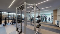 This fitness center offers a comprehensive workout environment organized across several distinct zones within a single open floor plan. The space is finished with a contemporary aesthetic, featuring sleek linear LED lighting along the ceiling, light gray walls, and striped multi-tone flooring that visually delineates workout areas.

The main floor houses an extensive array of cardio equipment, including Matrix treadmills, rowing machines, recumbent bikes, and ellipticals. A dedicated strength training area features a multi-functional cable and pull-up rig, a full rack of kettlebells, dumbbells, weight benches, and plate-loaded machines. Floor-to-ceiling mirrors expand the sense of space and allow users to monitor their form throughout workouts.

A glass-walled cycling studio with black-framed partitions provides a separate space for indoor cycling classes, offering visibility into the main floor. An adjoining lounge area features warm wood-paneled walls, pendant lighting, seating, built-in cabinetry, mini-refrigerators, and a wall-mounted television—providing a comfortable space to rest and recharge. Additional TVs are positioned throughout the main workout floor for entertainment during training sessions.