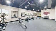 Located at 210 Reservation Way in Augusta, Georgia, this dedicated fitness center offers a well-appointed exercise space across a single open floor. The room is generously sized, providing ample space for a diverse range of workout equipment and free movement throughout.

The interior features an exposed industrial-style ceiling finished in dark paint, lending the space a modern, gym-appropriate aesthetic. Overhead, large ceiling fans ensure airflow and comfort during workouts, complemented by bright recessed and track lighting throughout. The flooring consists of durable gray carpet designed to support both cardio and strength training activities.

The exercise room is equipped with a variety of cardio machines, including treadmills, an elliptical trainer, a stationary bike, and a rowing machine. Strength training is supported by a cable machine, weight bench, and additional free-weight accessories. Exercise balls and yoga mats are also present for flexibility and core training.

Multiple framed wall mirrors are positioned throughout the space, allowing users to monitor their form, while windows along the walls bring in natural light and offer views of the surrounding outdoor greenery. Motivational artwork, including a large aerial stadium photograph, decorates the walls, adding character to the environment. Clearly marked emergency exits and accessible door access contribute to a safe and functional layout.